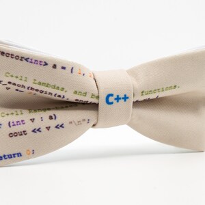 K&ouml;nnte beinhalten: Ein beiger Fliege mit wei&szlig;em Rand und einem C++-Logo in Blau. Die Fliege ist mit C++-Code bedruckt.