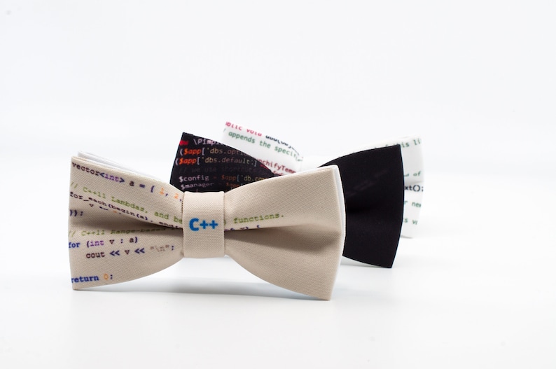 K&ouml;nnte beinhalten: Drei Fliegen mit einem Coding-Thema. Die Fliegen sind beige, schwarz und wei&szlig; mit Code bedruckt. Die beige Fliege hat den Text "C++" aufgedruckt.