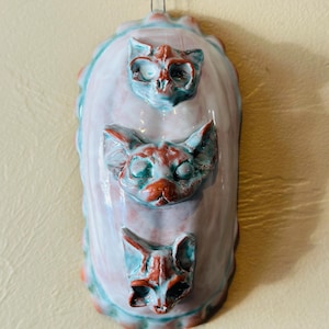 May include: A ceramic wall hanging with three cat faces sculpted in relief. The cat faces are in shades of red and white, with a light blue glaze. The background is white with a light blue glaze.