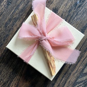 May include: A cream-colored gift box tied with a light pink chiffon ribbon. A piece of Palo Santo wood is placed on top of the box. The ribbon has frayed edges, and the box sits on a dark wood surface.