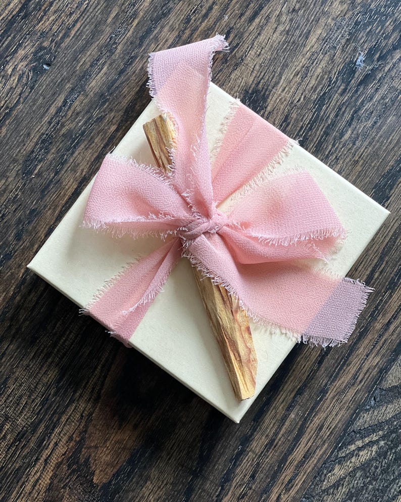 May include: A cream-colored gift box tied with a light pink chiffon ribbon. A piece of Palo Santo wood is placed on top of the box. The ribbon has frayed edges, and the box sits on a dark wood surface.