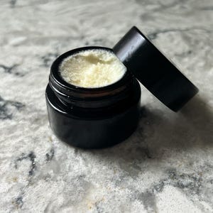 May include: A small, black jar with a matching lid, slightly ajar, revealing a creamy, pale yellow substance. The jar sits on a speckled, light-colored surface, suggesting a skincare or cosmetic product.