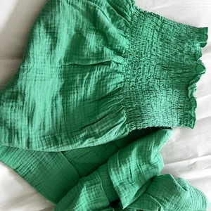 Peut inclure: Vêtement vert vif, possiblement un haut ou une robe, en tissu texturé. Il présente un col et des manches froncés et élastiqués. Le tissu a une texture froissée. L'article est présenté sur une surface blanche.