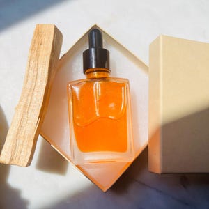 May include: A square glass bottle with a black dropper filled with an orange liquid, presented in a beige box. A piece of light brown wood is placed next to the box. The bottle is on a white surface.