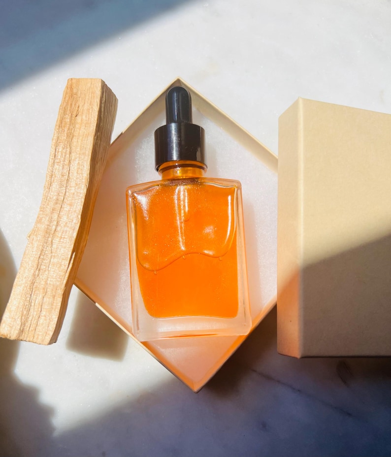 May include: A square glass bottle with a black dropper filled with an orange liquid, presented in a beige box. A piece of light brown wood is placed next to the box. The bottle is on a white surface.