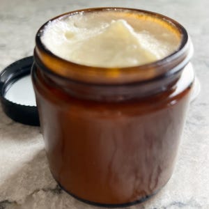 May include: A small, amber-colored glass jar filled with a creamy, off-white substance. The jar's black lid is placed next to it. The product appears to be a skincare cream or balm. The jar is sitting on a light-colored surface.