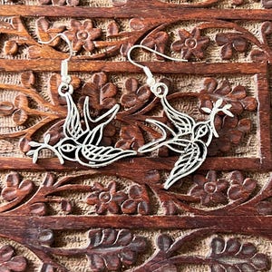 May include: A pair of silver-colored bird earrings. Each earring features a detailed cutout design of a bird carrying a sprig. The earrings are displayed on a carved wooden surface with floral patterns.
