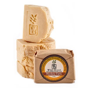 May include: Artisan soaps in a light brown hue, stacked with a carved design on top. A wrapped soap bar, labelled "Monkey Farts" and "Wild Grove", is visible in the foreground. The soap weighs 130g.