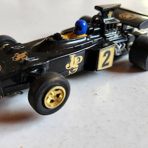 May include: Black and gold vintage toy race car with the number 2 on the side. The car has a blue helmeted driver and gold wheels. The car has the JP logo and other gold accents.