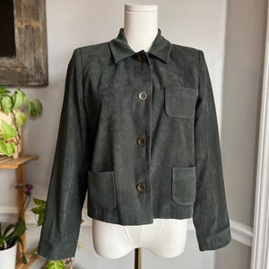 May include: A dark charcoal gray jacket with a soft, suede-like texture. It features a classic collar, long sleeves, and a front button closure with four dark brown buttons. The jacket includes one chest pocket and two lower pockets on the front.