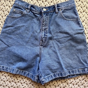 May include: High-waisted denim shorts in a medium blue wash featuring a classic five-pocket design, a metal button closure, a zippered fly, and reinforced copper-toned rivets at the pocket corners with visible stitching throughout the garment.