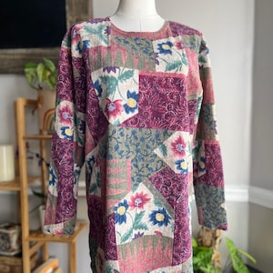 Puede incluir: Una blusa de manga larga con un diseño de patchwork que presenta varios patrones florales en tonos magenta intenso, verde salvia y crema. La prenda tiene un cuello redondo y una textura de tela marcada, exhibida sobre un torso de maniquí.