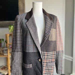 May include: A patchwork blazer in various shades of gray, brown, and black, displayed on a mannequin. The blazer features a mix of plaid, herringbone, and solid fabric panels, with a brown velvet collar and a single button closure. The blazer has two front pockets.