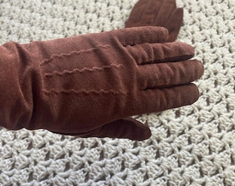 Op de afbeelding: Een paar bruine suède handschoenen met drie rijen decoratieve stiksels op de rug van de hand. De handschoenen hebben een zachte, matte textuur en vallen over de pols, met één handschoen op de voorgrond en de andere erachter.