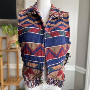Könnte beinhalten: Eine ärmellose Weste mit einem farbenfrohen geometrischen Muster. Die Weste hat einen blauen Hintergrund mit roten, grünen und beigen Zickzacklinien und Streifen. Der Kragen und der Saum sind mit Fransen versehen. Die West ist auf einer Schaufensterpuppe ausgestellt.