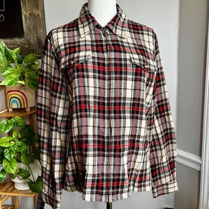 May include: A button-up plaid shirt in a classic pattern of red, black, and cream. The shirt has two chest pockets and a collared neckline. The fabric appears to be a medium-weight material, suitable for cooler weather.
