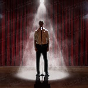 May include: A person stands on a stage under a spotlight, holding a microphone. Red curtains form the backdrop. The stage is illuminated with spotlights and a haze effect, creating a dramatic atmosphere. The person is wearing a white shirt and black pants.