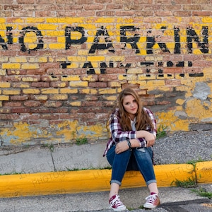 Puede incluir: Una joven con una camisa a cuadros y pantalones vaqueros azules se sienta en una acera frente a una pared de ladrillo con pintura amarilla. La pared tiene las palabras "NO PARKING ANY TIME" pintadas en ella.