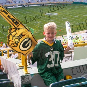 May include: A young boy wearing a green and white jersey with the number 30 on it, is holding a foam finger that says "#1 Fan" and a green and white foam tube. The boy is sitting in a stadium seat with a green and yellow railing in front of him. The background is a green football field.