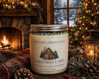 May include: A clear glass jar candle with a silver metal lid and a white label featuring a log cabin illustration, green pine trees, and the text CLIP CLOP CANDLE SHOP CHRISTMAS CABIN HAND POURED IN EAST TENNESSEE 100% SOY WAX, tied with green and white twine.