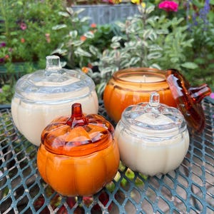 Può includere: Quattro portacandele in vetro a forma di zucca con coperchi. Due sono arancioni, uno è trasparente e uno è color crema chiaro. Le candele sono su un tavolo di metallo verde con sfondo giardino.