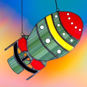 May include: Stained glass rocket ship suncatcher with red, yellow, green, and white accents. The rocket has a red nose with white dots, a yellow stripe, and green body. Black metal outlines the design. Includes a hanging loop.
