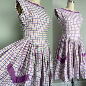 May include: A vintage 1950s style dress with a purple and white checkered pattern. The dress has a fitted bodice and a full skirt with pockets. The dress is sleeveless and has a scoop neckline.