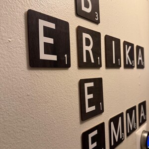 May include: Wooden Scrabble tiles spelling out the names ERIKA, EMMA, and B. The tiles are black with white letters and point values. The tiles are mounted on a light beige wall.