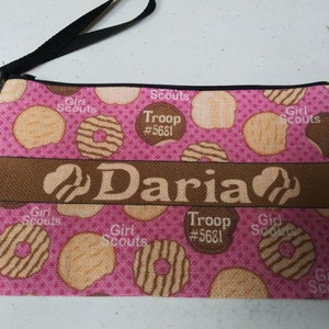 May include: Pink zippered pouch with a brown strap. The pouch is decorated with a pattern of Girl Scout cookies and the text "Girl Scouts", "Troop #5681", and "Daria".