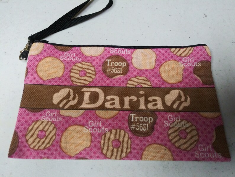 May include: Pink zippered pouch with a brown strap. The pouch is decorated with a pattern of Girl Scout cookies and the text "Girl Scouts", "Troop #5681", and "Daria".