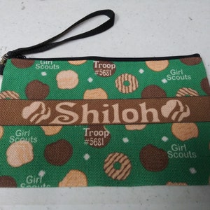 May include: Green zippered pouch with a brown strap. The pouch has a green background with a pattern of brown and beige circles. The text "Girl Scouts" and "Troop #5681" is printed on the pouch. The name "Shiloh" is printed in a brown banner across the middle of the pouch.