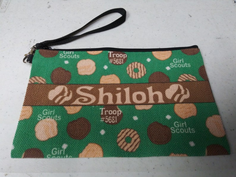 May include: Green zippered pouch with a brown strap. The pouch has a green background with a pattern of brown and beige circles. The text "Girl Scouts" and "Troop #5681" is printed on the pouch. The name "Shiloh" is printed in a brown banner across the middle of the pouch.