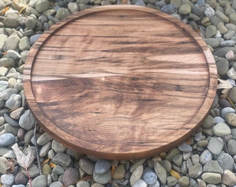 May include: A large, round wooden serving tray with a natural wood finish. The tray is made of dark wood with a grain pattern. The tray is sitting on a bed of small, gray stones.