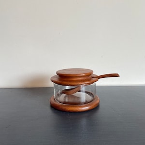 May include: A wooden lidded glass jar with a wooden spoon inside. The jar is made of clear glass and has a wooden base and lid. The spoon is made of wood and has a curved handle.
