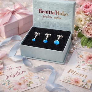 May include: A set of three silver stud earrings with blue opal-like stones, presented in a light blue gift box. The box is open, revealing the earrings on a black velvet interior. The box has the text "Benitta Moko fashion online".