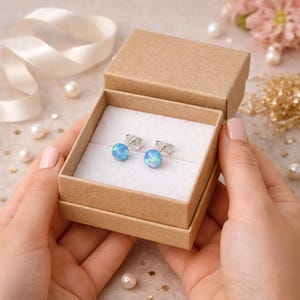 Könnte beinhalten: Ein Paar runde, leuchtend blaue Ohrstecker mit schimmernder Oberfläche, gefasst in silberfarbenem Metall. Die Ohrringe werden in einer kleinen, quadratischen Geschenkbox aus hellbraunem Karton mit weißer Polstereinlage präsentiert.