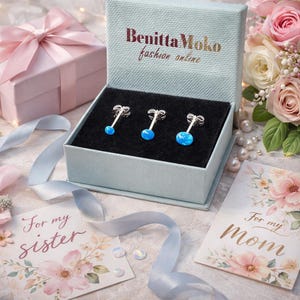 May include: A light blue jewelry box with the text BenittaMoko fashion online printed on the lid. Inside, three silver stud earrings with round, bright blue opal-like stones are set against a black velvet insert. The box is open.