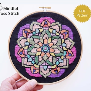 May include: A cross-stitch mandala design in a wooden embroidery hoop. The design features a symmetrical pattern with shades of pink, purple, green, orange, and blue on a black background. The text "Mindful Cross Stitch" and "PDF Pattern" are also visible.