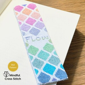 May include: A rectangular cross-stitch bookmark featuring a gradient pattern of diamond shapes in shades of pink, orange, purple, green, and blue on a white fabric background. The word FLOW is stitched in light green letters in the center.