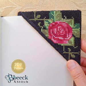 May include: A black fabric bookmark with a cross-stitch design of a red rose with green leaves. The bookmark is folded over the corner of a white notebook. The text "PDF Pattern" is on a gold circle sticker on the notebook.