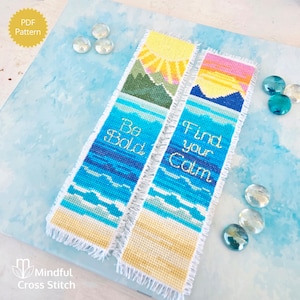 May include: Two cross-stitch bookmarks with beach and mountain scenes. One says "Be Bold" and the other says "Find your Calm". The bookmarks have a yellow sun, blue water, and green mountains. The bookmarks are on a blue surface with clear glass beads.