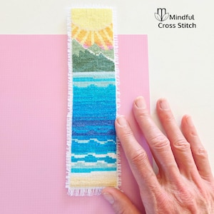 May include: A handmade cross-stitch bookmark featuring a beach scene with a yellow sun, green mountains, and blue ocean. The bookmark has a frayed edge and is held against a pink background. The text "Mindful Cross Stitch" is in the upper right corner.