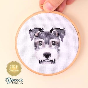 Può includere: Uno schema a punto croce di un cane schnauzer miniatura in un telaio da ricamo in legno. Lo schema è disponibile per il download in PDF.