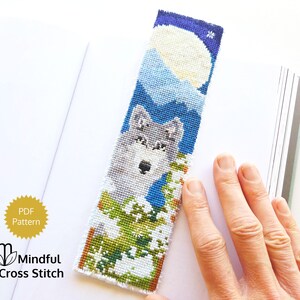 May include: A cross-stitched bookmark featuring a wolf in a snowy landscape under a full moon. The bookmark has a blue background with white and gray details. The text "Mindful Cross Stitch" is visible.