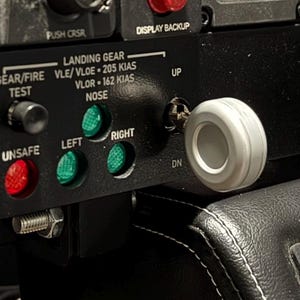 May include: Close-up of an aircraft control panel with various buttons and switches. The panel is black with white text and includes labels like "PITOT HEAT" and "LANDING GEAR." A white, circular handle is prominent, and there are green and red indicator lights.