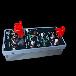 May include: A rectangular control panel with multiple toggle switches and indicator lights. The panel has a silver frame and a black face with white lettering. Two red safety covers are attached to the panel. The panel includes the text "AVIONICS" and "Deslen".