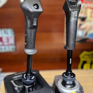 May include: Two black flight simulator control sticks with textured grips and red button tips. One stick is mounted in a black box, the other on a silver base. The sticks have various buttons and labels.