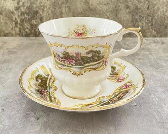 May include: A cream-colored porcelain teacup and saucer set featuring a decorative gold filigree border, a landscape scene with a stone tower, and small floral accents. The cup has a pedestal base and a curved handle with gold trim detailing.