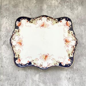 May include: A rectangular ceramic serving tray with scalloped edges, featuring a deep navy blue border and a cream-colored center. The surface is decorated with ornate gold scrollwork and delicate floral patterns in shades of pink and orange.
