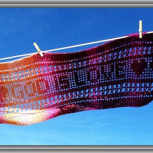 May include: A rectangular crochet scarf featuring a gradient of orange, pink, and purple yarn. The scarf displays the text "GOD IS LOVE" in a block-style pattern, accented by heart shapes, and is suspended horizontally by three wooden clothespins.